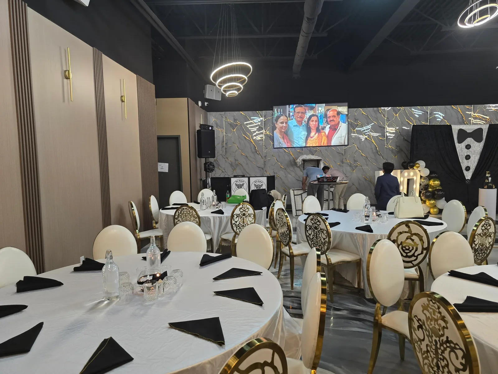 Banquet hall set for an event at Rawaayt in Ellerslie, Edmonton — round tables with white linen, black napkins and gold-framed chairs under ring chandeliers