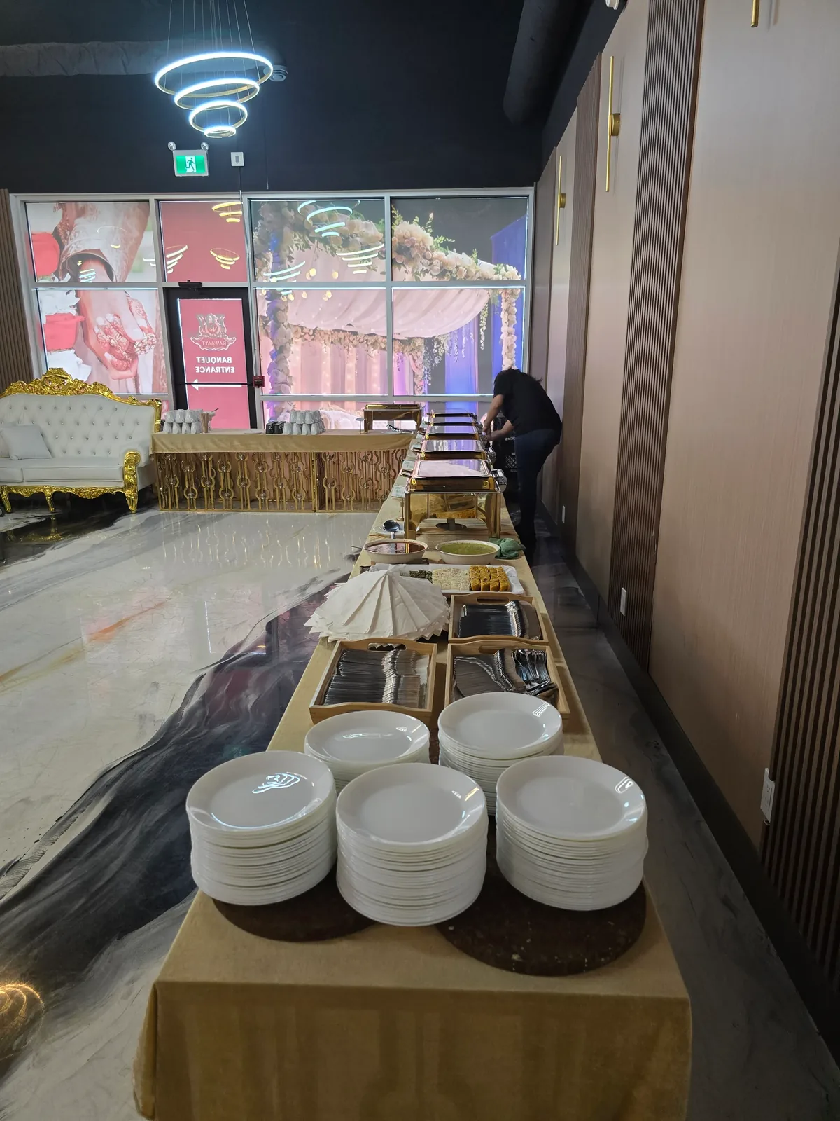 Buffet line set for service at the Rawaayt banquet hall in Edmonton — stacked plates, cutlery trays and gold chafing dishes along a skirted table as staff plate the food