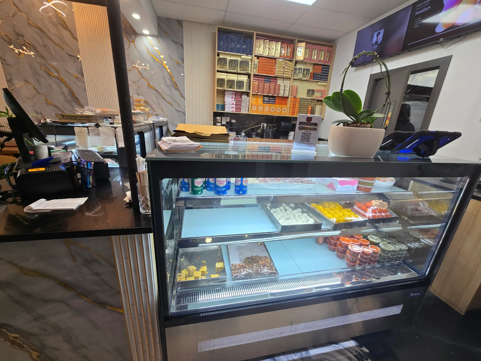 Indian sweets display counter with rasgulla, boondi ladoo and gulab jamun beside a wall of mithai gift boxes at Rawaayt, Ellerslie Edmonton